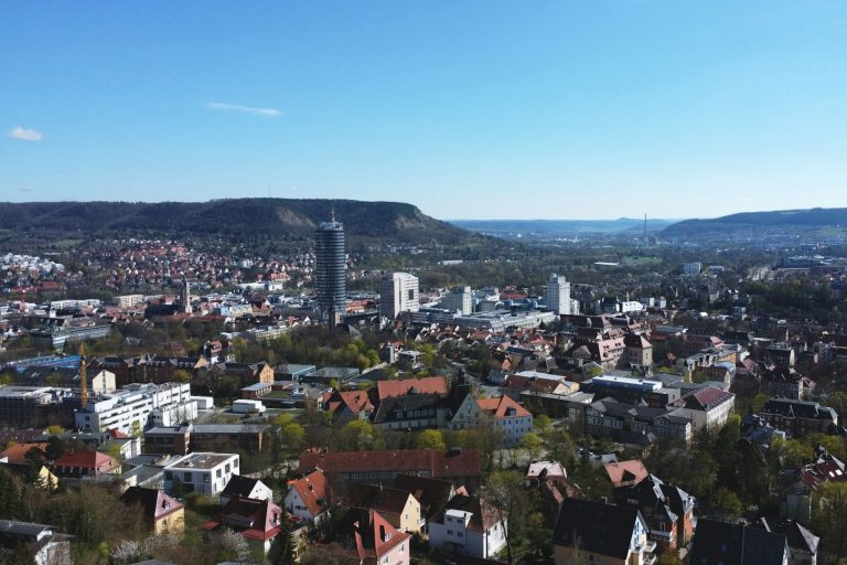 Blick auf eine Stadtlandschaft  von Jena mit Bergen und modernen Gebäuden im Hintergrund.