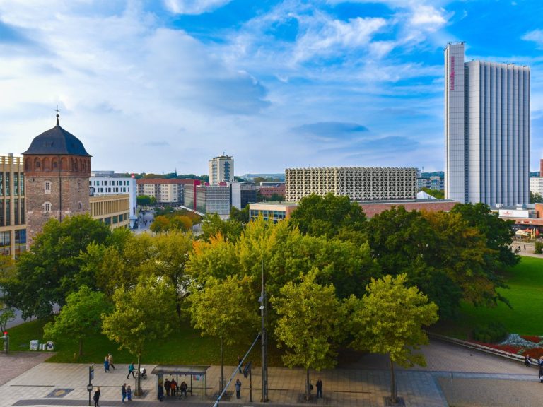 Stadtansicht Chemnitz mit modernen Gebäuden und Bäumen, unter blauem Himmel mit Wolken.