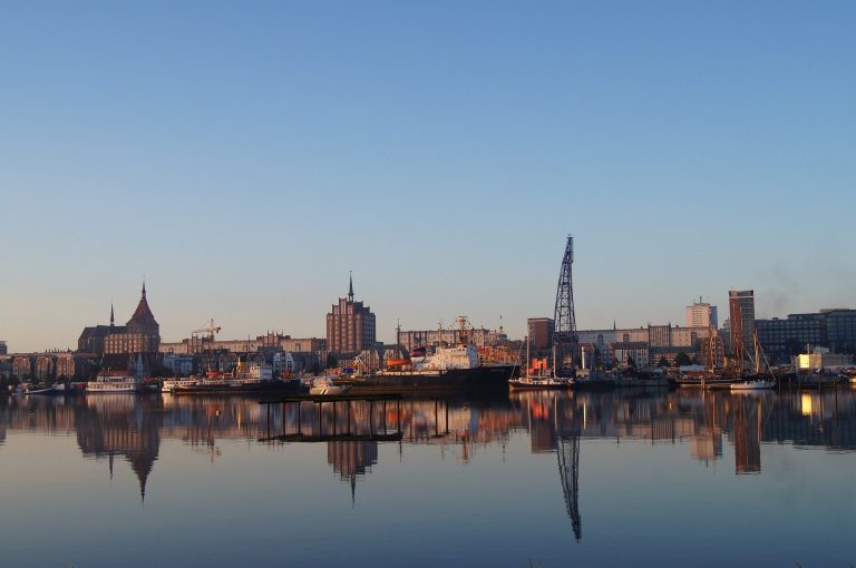 Stadtansicht mit Gebäuden und Hafen Rostock bei Sonnenaufgang, reflektiert im Wasser.