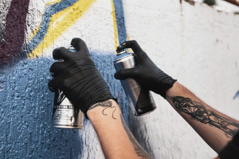 Hände mit Sprühdosen, die an einer Wand graffitiartige Kunst auftragen.