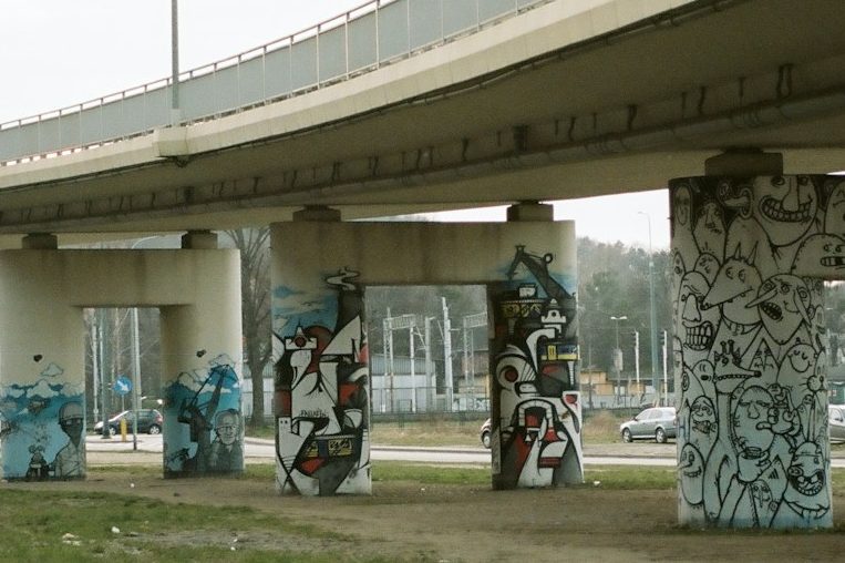 Graffiti-verzierte Pfeiler unter einer Brücke, umgeben von städtischem Umfeld.