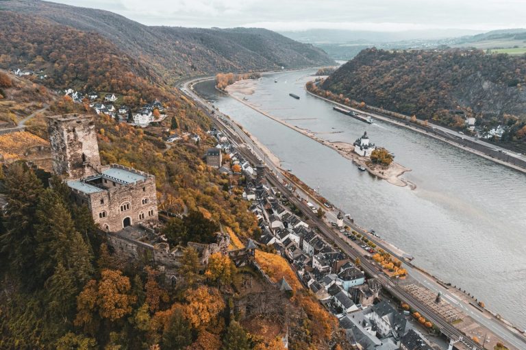 Landschaft mit Fluss, Herbstbäumen und einer historischen Burg auf einem Hügel.