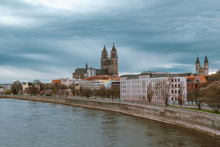 Stadtansicht mit Fluss, Häusern und einer Kirche im Hintergrund unter bewölktem Himmel.