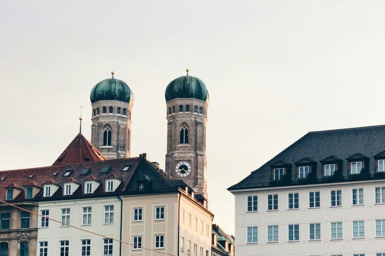 Zwei Türme mit grünen Kuppeln vor einem hellen Himmel über historischen Gebäuden.