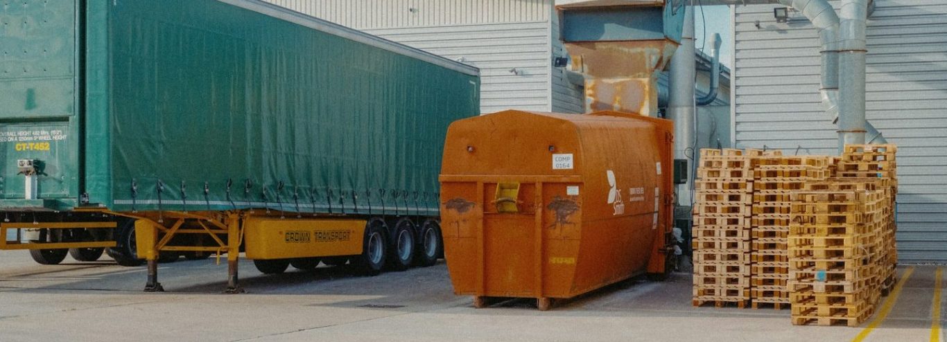 Speditionsdelikte | Transportdiebstahl Berlin Lagerbereich mit einem grünen Lkw-Anhänger, einem orangefarbenen Container und Holzpaletten.