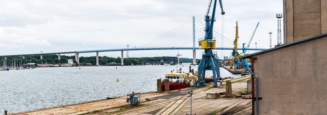 Blick auf einen Hafen mit Kränen, Booten und einer Brücke im Hintergrund.