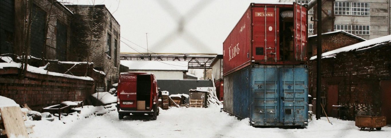 Lagerhalle mit verschneitem Boden und Lagercontainern, umgeben von Gebäuden.
