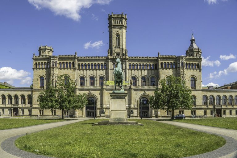 Historisches Gebäude mit Turm und Statue, umgeben von Wiesen und blauem Himmel.