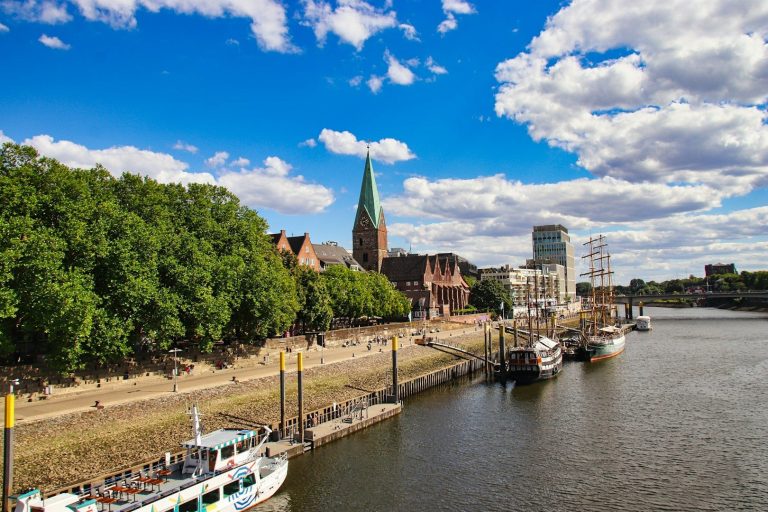 Blick auf einen Fluss mit Booten, Uferpromenade und einer Kirche im Hintergrund in Bremen.