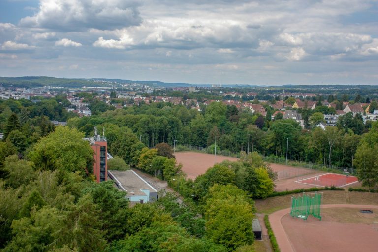 Landschaft mit Tennisplätzen und Wohngebieten, umgeben von Bäumen und Wolken.