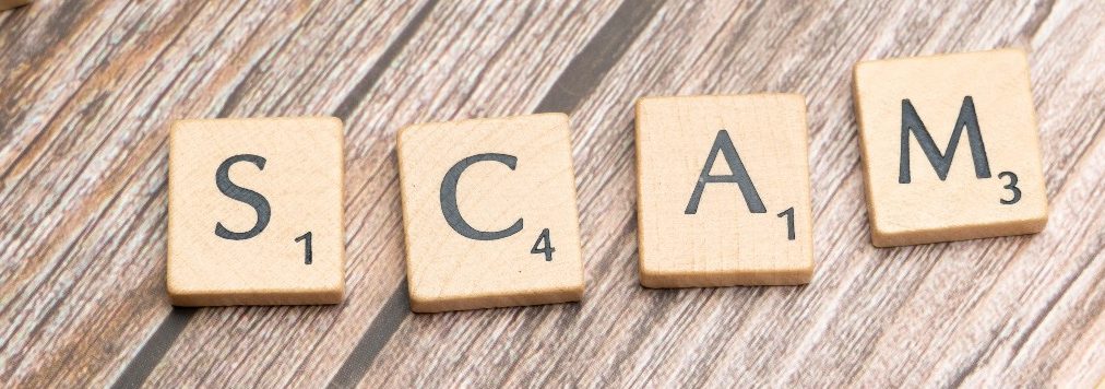 Buchstabenplättchen spellend das Wort „SCAM“ auf einem Holzuntergrund.